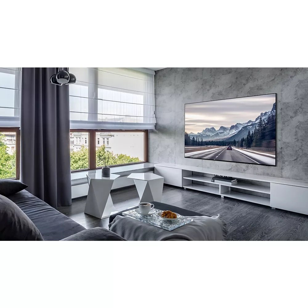 Un salon moderne présente l'élégant téléviseur SAMSUNG TQ77S95D avec une superbe scène de montagne, complétée par de grandes fenêtres qui laissent entrer la lumière naturelle et une table basse minimaliste améliorant l'ambiance tranquille.