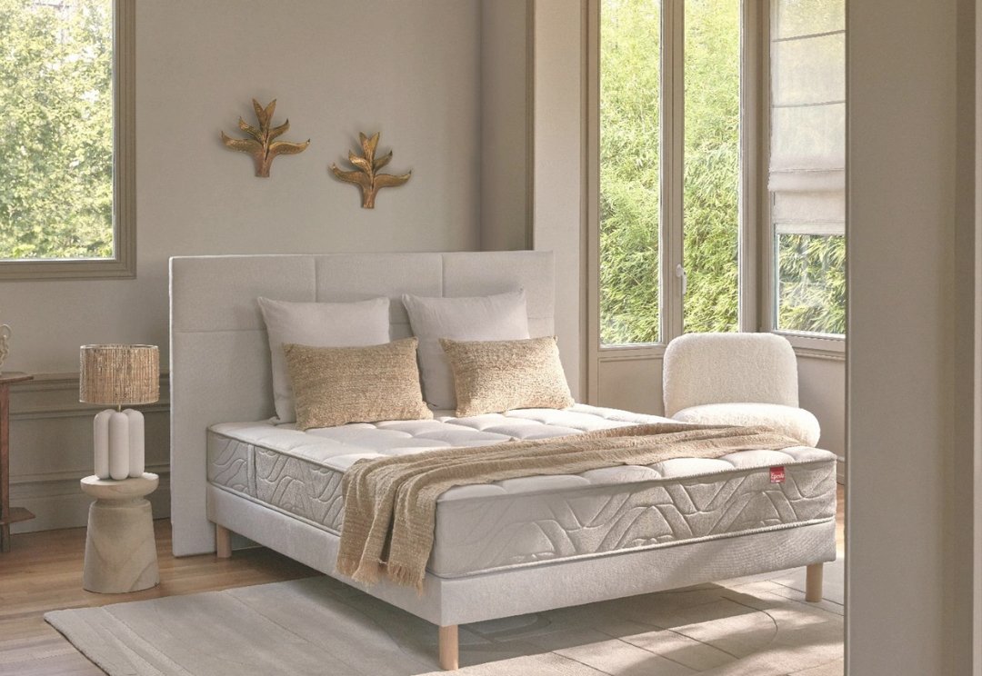 Chambre à coucher lumineuse avec un lit EPEDA EMBRUN blanc, des oreillers beiges, une chaise, de grandes fenêtres et un art mural en forme de feuille.