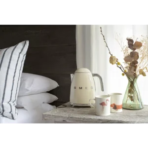 La bouilloire SMEG KLF03BLMEU est posée sur une table de chevet rustique, à côté d'un oreiller rayé, avec des tasses à motif animalier et des fleurs séchées.