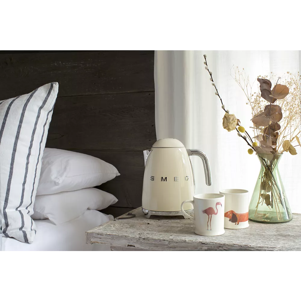La bouilloire SMEG KLF03BLMEU est posée sur une table de chevet rustique, à côté d'un oreiller rayé, avec des tasses à motif animalier et des fleurs séchées.