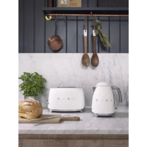 La bouilloire SMEG KLF03BLMEU et le grille-pain blanc SMEG sont posés sur un comptoir en marbre, avec du pain, du basilic et des ustensiles en bois suspendus au-dessus.