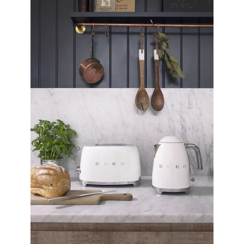 La bouilloire SMEG KLF03BLMEU et le grille-pain blanc SMEG sont posés sur un comptoir en marbre, avec du pain, du basilic et des ustensiles en bois suspendus au-dessus.