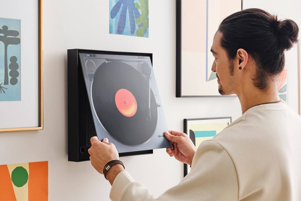 Personne plaçant un disque vinyle dans un tourne-disque mural, entourée d'œuvres d'art encadrées et d'une élégante barre de son SAMSUNG HW-LS60D.