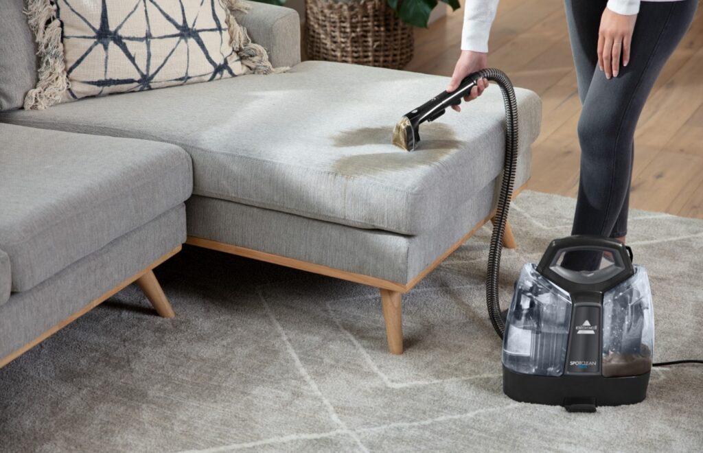 Une personne utilise un BISSELL SPOTCLEAN PET SELECT 37288 pour nettoyer méticuleusement un canapé gris sur un tapis dans un salon confortable.