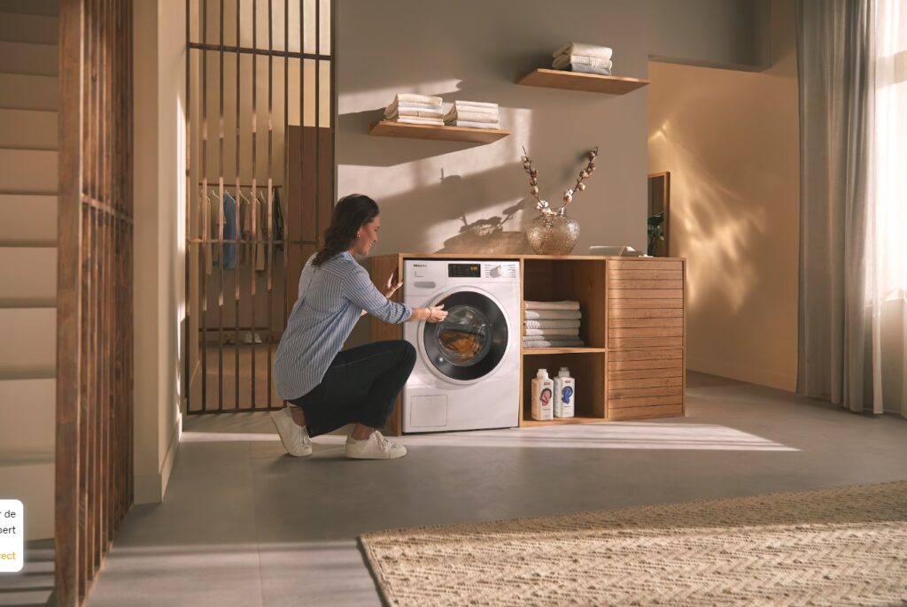 Personne agenouillée pour charger une machine à laver MIELE WWD164 WCS dans une buanderie moderne avec étagères, serviettes et produits de nettoyage.