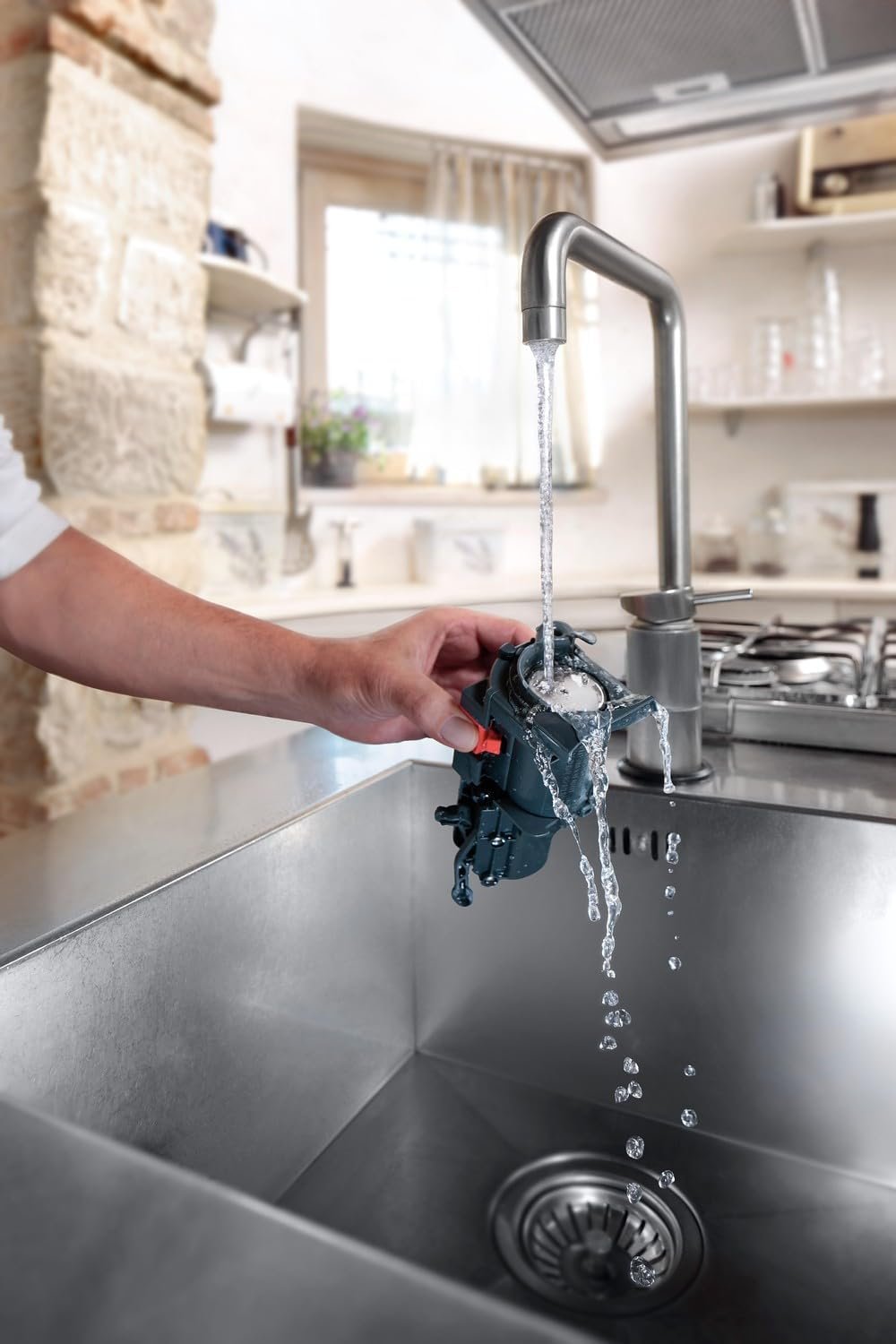 Une personne rince un petit appareil mécanique étiqueté DELONGHI ECAM293.52B MAGNIFICA sous un robinet de cuisine en marche dans une cuisine moderne avec un évier en acier inoxydable.