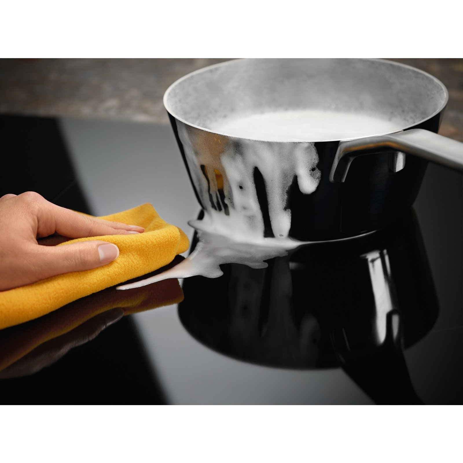 Nettoyage à la main du lait renversé sur une cuisinière ELECTROLUX EIV63343 avec un chiffon jaune, tandis que le lait continue de bouillir dans une casserole.