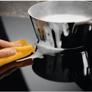 Une main avec un chiffon jaune essuie la cuisinière d'un ELECTROLUX IDE6342KC où une casserole de lait bouillant a débordé.