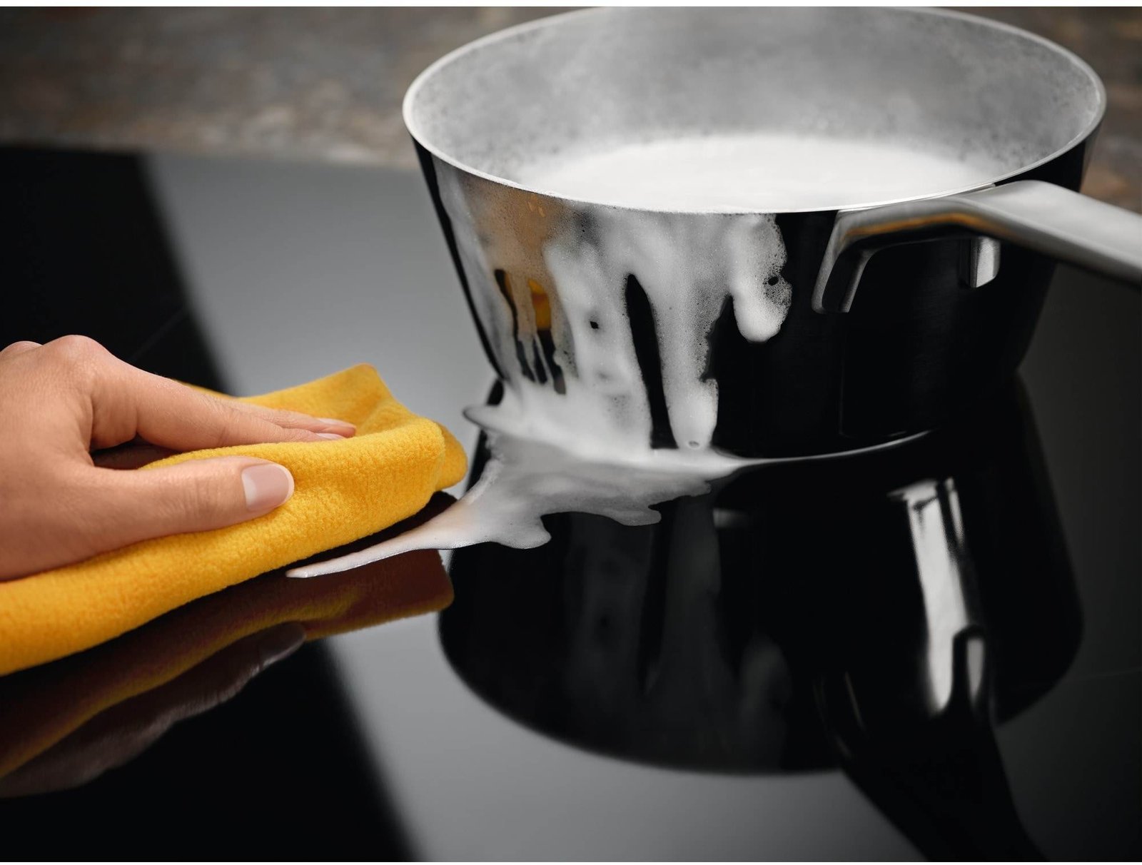Une main avec un chiffon jaune essuie la cuisinière d'un ELECTROLUX IDE6342KC où une casserole de lait bouillant a débordé.
