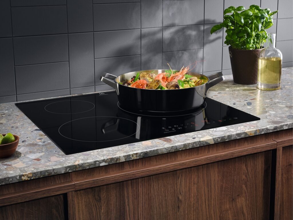 Une poêle contenant des aliments mijote sur la table de cuisson à induction moderne ELECTROLUX IDE6342KC, posée sur un comptoir de cuisine élégant à côté d'une plante en pot et d'une bouteille d'huile.
