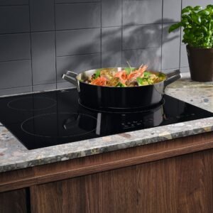 Une poêle contenant des aliments mijote sur la table de cuisson à induction moderne ELECTROLUX IDE6342KC, posée sur un comptoir de cuisine élégant à côté d'une plante en pot et d'une bouteille d'huile.