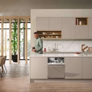 Un homme se tient debout dans une cuisine moderne, ouvrant son lave-vaisselle MIELE G 5150 VI. L'espace épuré est complété par des armoires en bois et un coin repas lumineux avec une grande fenêtre, créant une atmosphère accueillante.