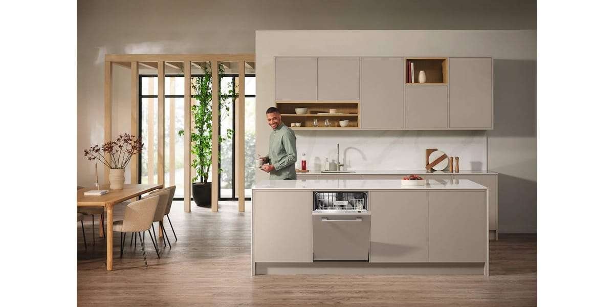 Un homme se tient debout dans une cuisine moderne, ouvrant son lave-vaisselle MIELE G 5150 VI. L'espace épuré est complété par des armoires en bois et un coin repas lumineux avec une grande fenêtre, créant une atmosphère accueillante.
