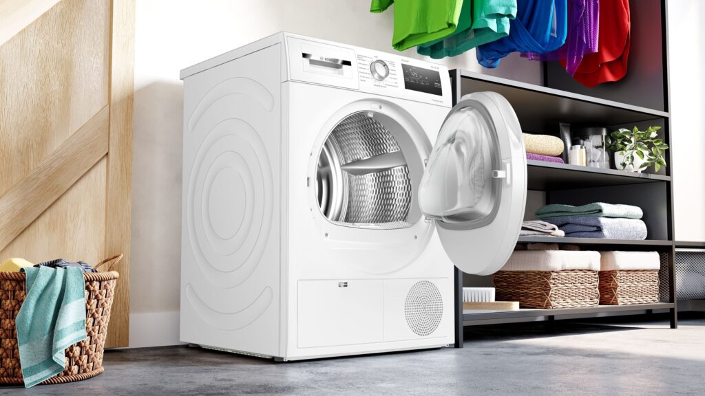 Un sèche-linge moderne BOSCH WTH8300MFR se trouve dans une buanderie avec sa porte ouverte, flanqué de vêtements soigneusement pliés sur des étagères et d'un panier de linge à proximité.