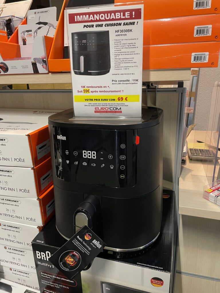 Une élégante friteuse à air Braun, modèle HF3030BK, occupe le devant de la scène sous un panneau promotionnel, ce qui en fait une idée cadeau idéale pour les amateurs de cuisine dans ce cadre de magasin accueillant.