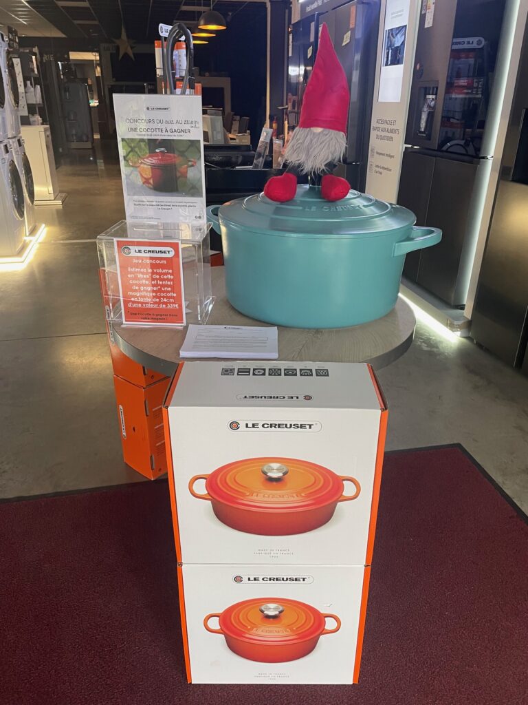 La vitrine du magasin Le Creuset présente une grande marmite turquoise, des boîtes élégamment empilées et un gnome décoratif fantaisiste portant un chapeau rouge. Gardez un œil sur notre prochain JEU CONCOURS pour avoir une chance de gagner l'une de ces pièces emblématiques !