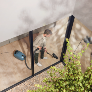 Vue d'en haut à travers une grande fenêtre en verre, une personne utilisant l'aspirateur Miele GUARD M1 FLEX sur un plancher en bois dans une pièce moderne incarne une propreté irréprochable.