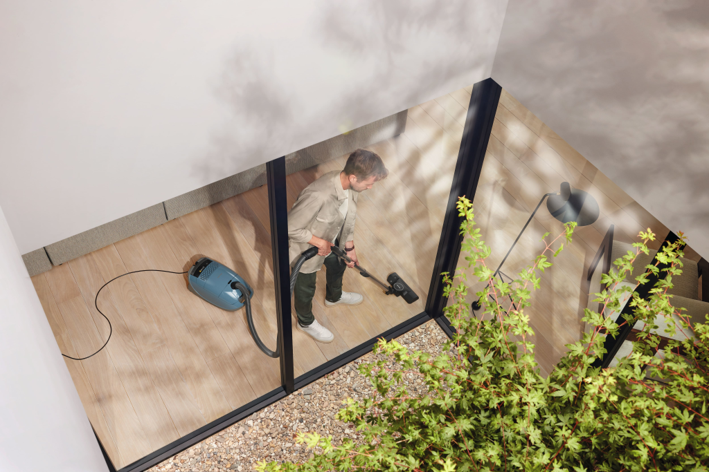 Vue d'en haut à travers une grande fenêtre en verre, une personne utilisant l'aspirateur Miele GUARD M1 FLEX sur un plancher en bois dans une pièce moderne incarne une propreté irréprochable.