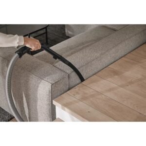 Une personne nettoie méticuleusement le sol à côté d'un canapé gris à l'aide de l'aspirateur Miele GUARD M1 FLEX.