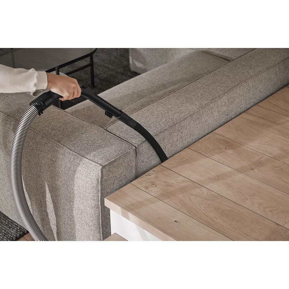 Une personne nettoie méticuleusement le sol à côté d'un canapé gris à l'aide de l'aspirateur Miele GUARD M1 FLEX.