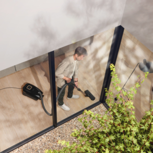À l'aide d'un aspirateur Miele GUARD L1 AllFloor Noir, quelqu'un passe l'aspirateur sur un sol en bois dans une pièce moderne aux parois de verre, avec une végétation luxuriante à l'extérieur.