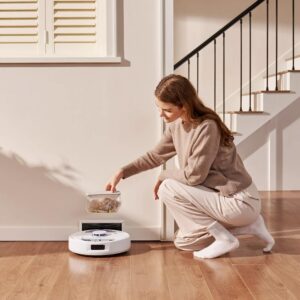 À côté de l'escalier, une femme accroupie sur un plancher en bois, plaçant soigneusement les débris dans l'aspirateur robot NARWAL FREO X PLUS.