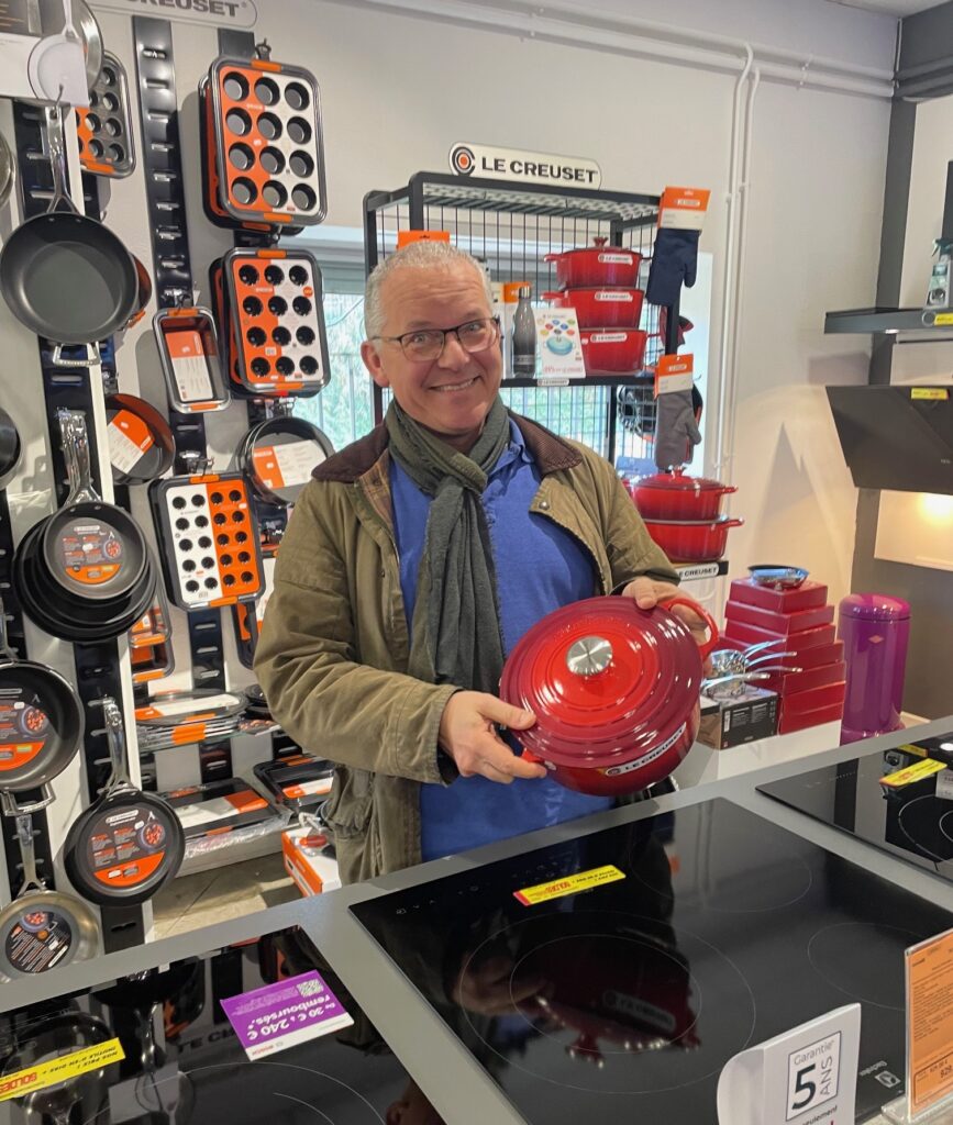 Dans un magasin de cuisine animé, un homme en manteau tient fièrement un couvercle d'ustensile de cuisine rouge LE CREUSET au milieu d'un assortiment d'articles de cuisine, se sentant comme le gagnant du jeu parmi LES GÉANTS de l'artisanat culinaire.