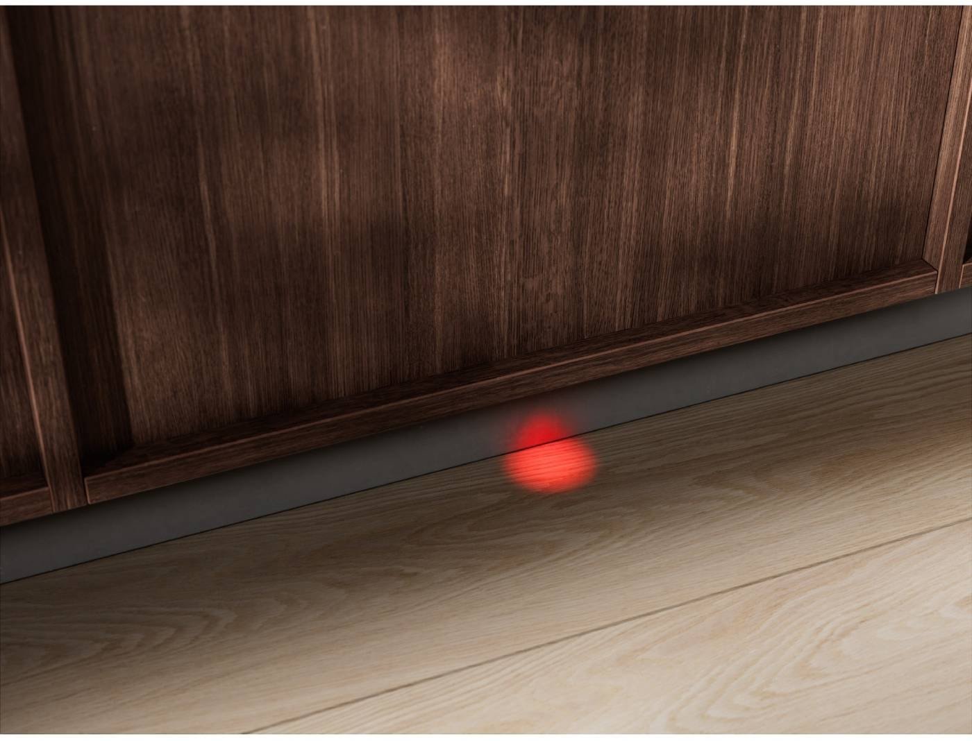 Un point laser rouge danse sur le plancher en bois devant un meuble, projetant des reflets subtils près des lignes épurées d'un lave-vaisselle ELECTROLUX EES48320L.