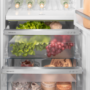 Le réfrigérateur LIEBHERR CBNSFC57Vi-22 présente un intérieur bien organisé, avec des légumes, des raisins, des bouteilles, des bocaux et des fromages emballés soigneusement disposés sur ses étagères.