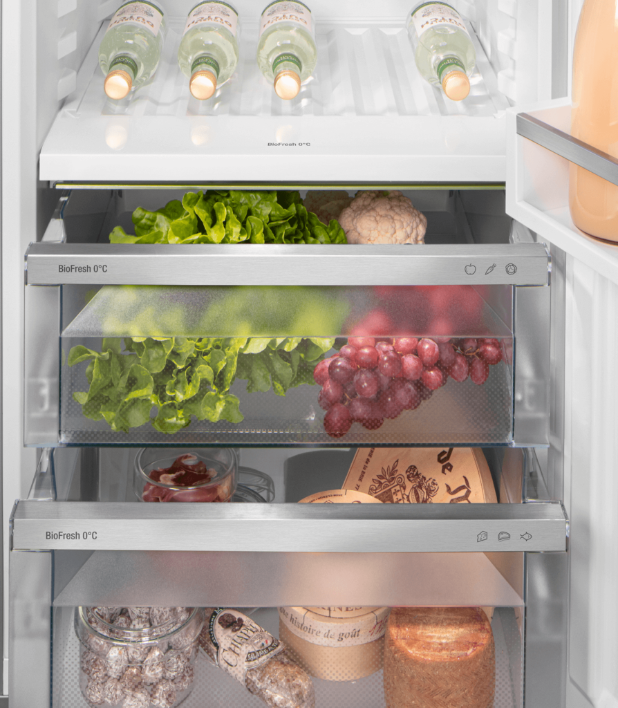 Le réfrigérateur LIEBHERR CBNSFC57Vi-22 présente un intérieur bien organisé, avec des légumes, des raisins, des bouteilles, des bocaux et des fromages emballés soigneusement disposés sur ses étagères.