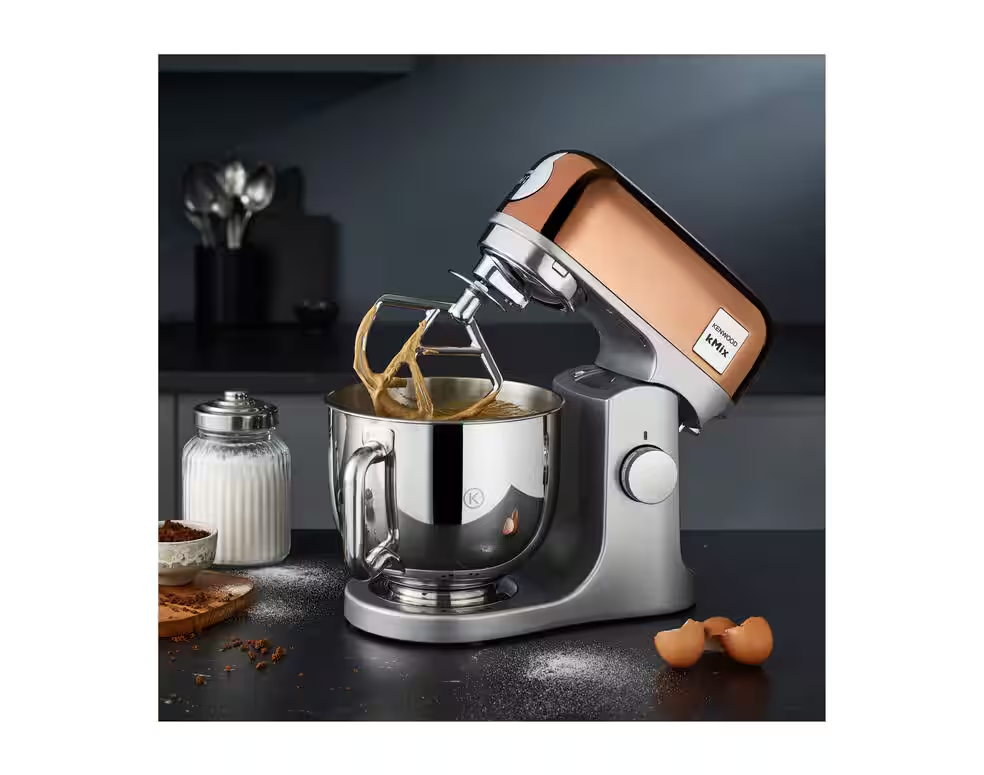 L'élégant batteur sur socle en cuivre MAGIMIX KMX760GD avec un bol en acier inoxydable se place élégamment sur le comptoir de la cuisine au milieu de divers ingrédients de cuisson.