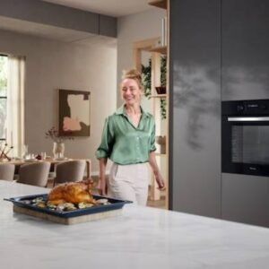 Une femme souriante vêtue d'un chemisier vert s'approche d'une table avec un plat parfaitement rôti, préparé dans sa cuisine moderne MIELE H2766BP NOIR.