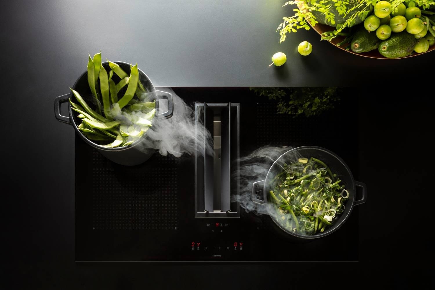 Vue de dessus de l'élégante cuisinière noire FALMEC QUANTUM3421 EASY avec des pots fumants de légumes-feuilles et un bol de fruits au citron vert à proximité, soulignant sa fonctionnalité facile.
