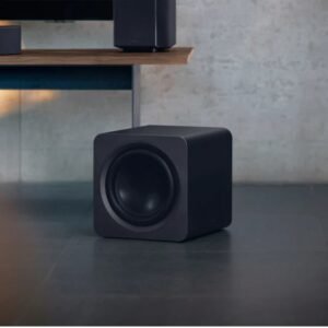 Un caisson de basses noir en forme de cube SAMSUNG HW-Q995D (Copie) est posé sur un sol carrelé gris près d'une étagère en bois.