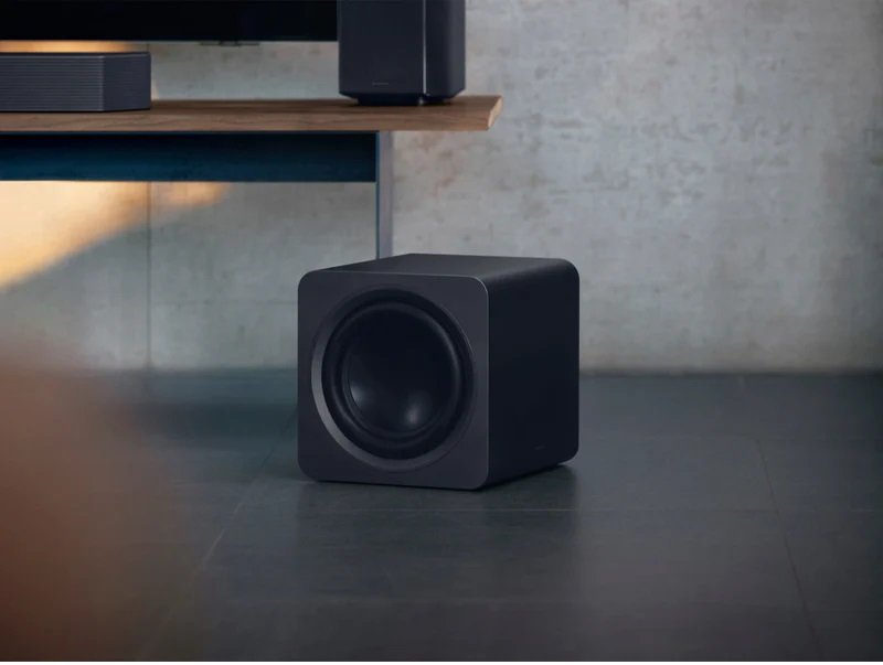Un caisson de basses noir en forme de cube SAMSUNG HW-Q995D (Copie) est posé sur un sol carrelé gris près d'une étagère en bois.