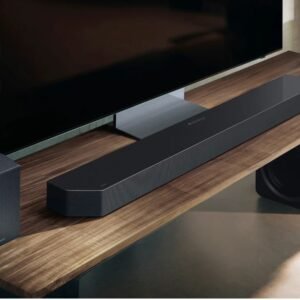 Une barre de son noire SAMSUNG HW-Q995D, des haut-parleurs et un caisson de basses sont placés sur un support TV en bois sous un téléviseur à écran plat.