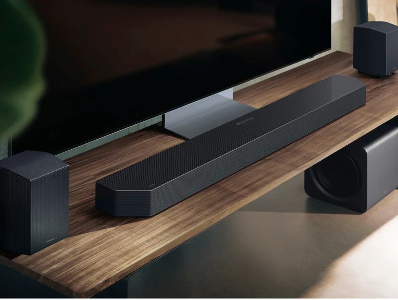 Une barre de son noire SAMSUNG HW-Q995D, des haut-parleurs et un caisson de basses sont placés sur un support TV en bois sous un téléviseur à écran plat.