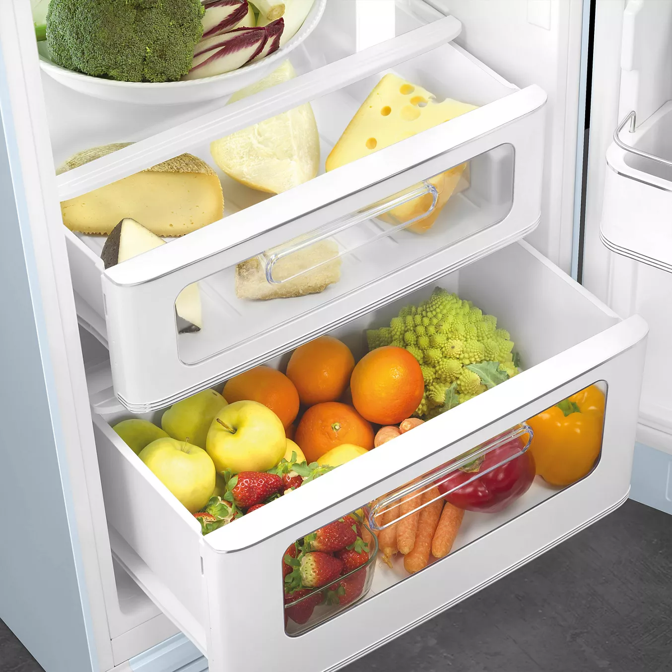 En ouvrant le réfrigérateur SMEG FAB30RPB5, vous trouverez des tiroirs remplis de fromage, d'oranges, de pommes et de légumes, ainsi que des brocolis sur une étagère.