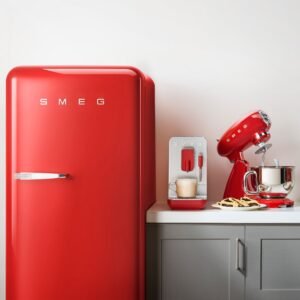 Le réfrigérateur rétro rouge SMEG FAB28RRD6 se dresse sur un comptoir blanc avec une machine à café et un mixeur assortis, des biscuits à côté d'eux, posés contre un mur clair.