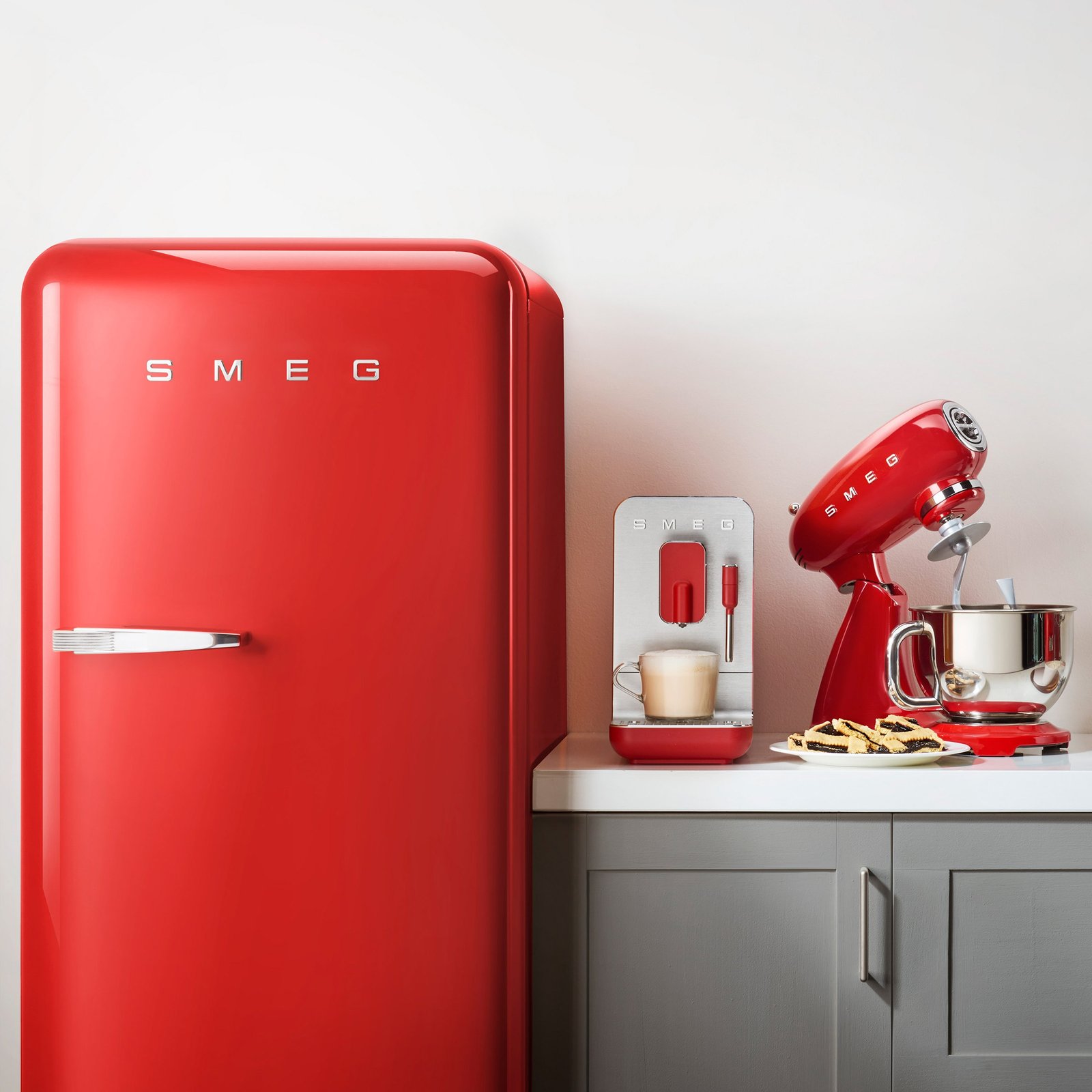 Le réfrigérateur rétro rouge SMEG FAB28RRD6 se dresse sur un comptoir blanc avec une machine à café et un mixeur assortis, des biscuits à côté d'eux, posés contre un mur clair.