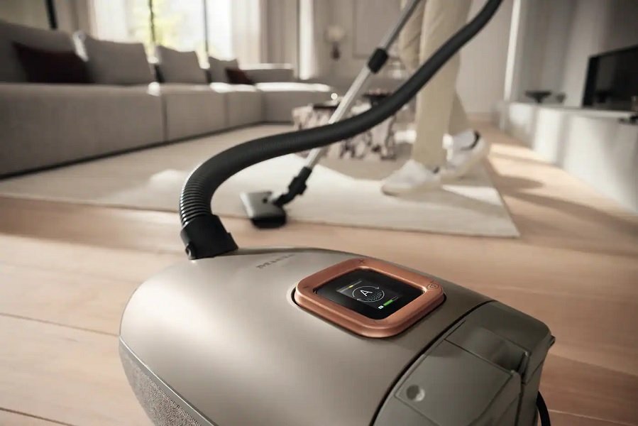 Gros plan d'un modèle de nouveaux aspirateurs sur un plancher en bois, avec une personne passant l'aspirateur sur le tapis du salon en arrière-plan.