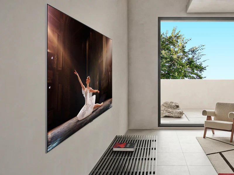 Dans le salon chic du LG OLED55G5 GALLERY DESIGN de 2025, un téléviseur OLED reproduit les mouvements élégants d'une danseuse. Une chaise et un livre sont posés au sol à proximité, avec en toile de fond la tranquillité des arbres.