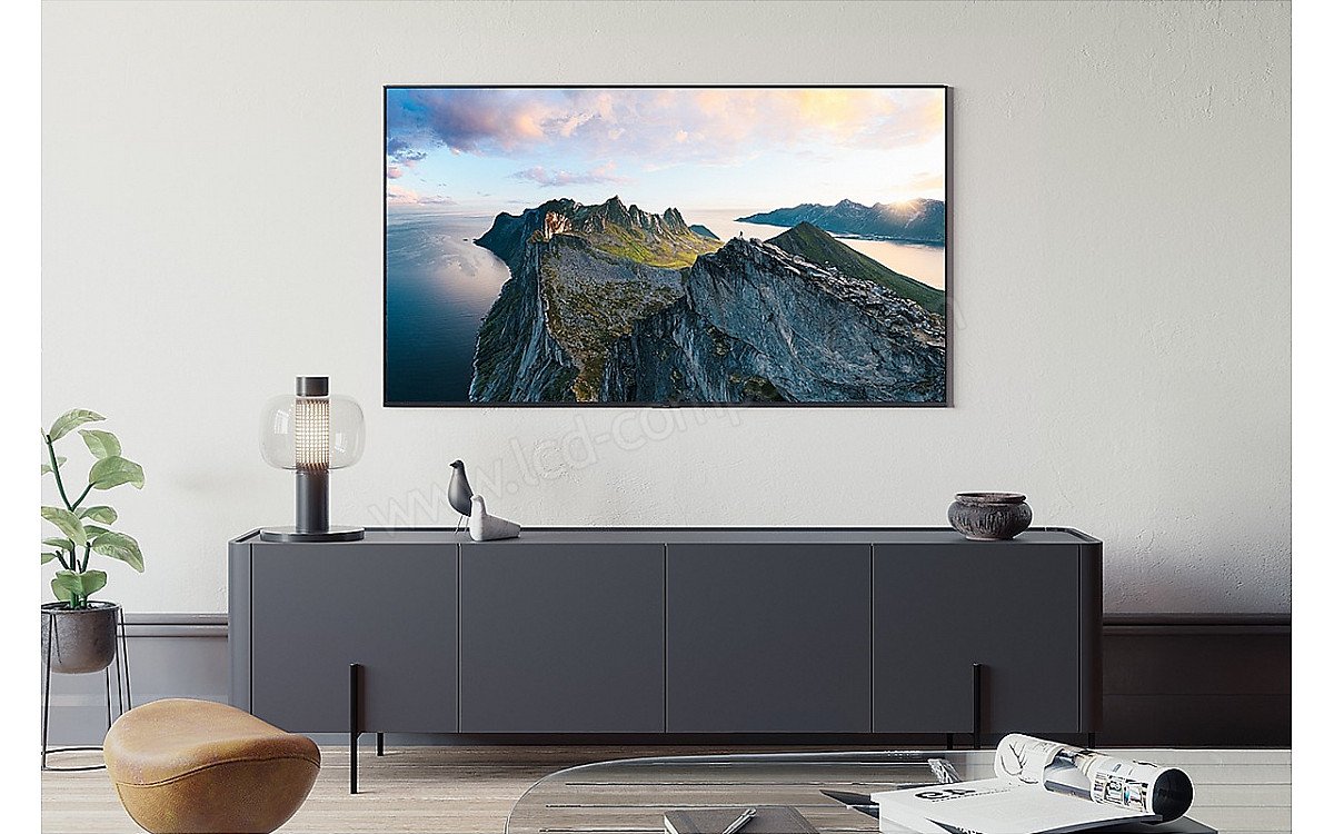 Un salon moderne avec une armoire sombre, une décoration de bon goût et une grande photo de montagne est rehaussé par l'élégant téléviseur SAMSUNG TQ100QN80F accroché au mur, ajoutant de la sophistication à l'espace.