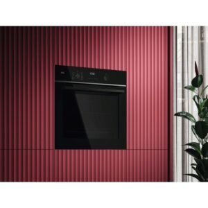 Le four encastrable AEG TA5PB53XAB noir est installé dans un mur texturé rose, avec une plante et un rideau blanc à droite.