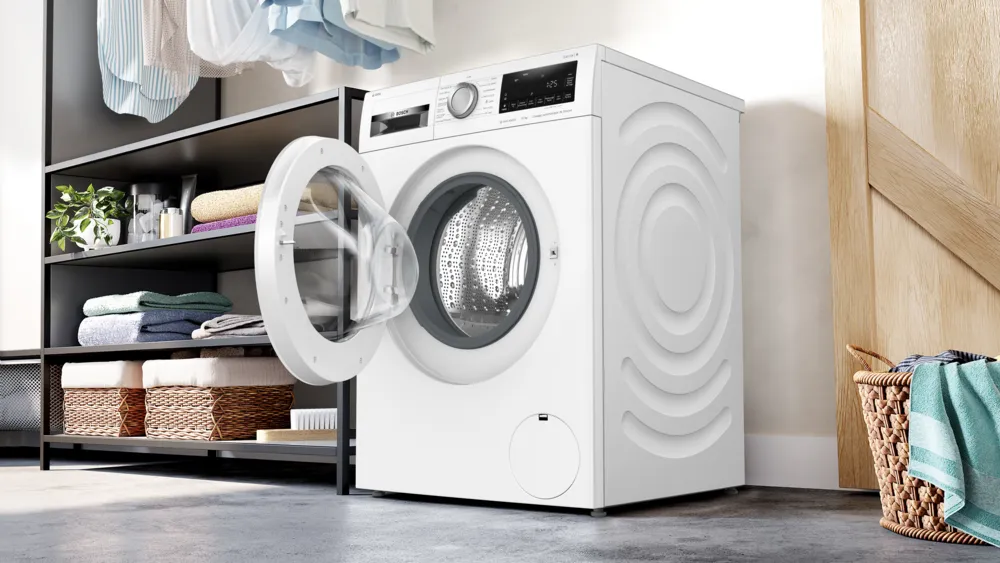 Le lave-linge BOSCH WGG254FAFR, porte ouverte, est présenté dans une buanderie moderne à côté d'étagères empilées avec des serviettes soigneusement pliées.