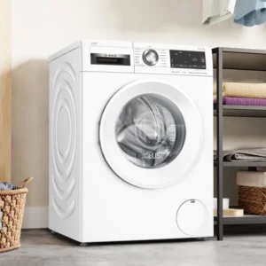Un lave-linge BOSCH WGG254FAFR se trouve à côté d'un panier à linge, avec des étagères au-dessus soigneusement rangées avec des serviettes pliées et des paniers.