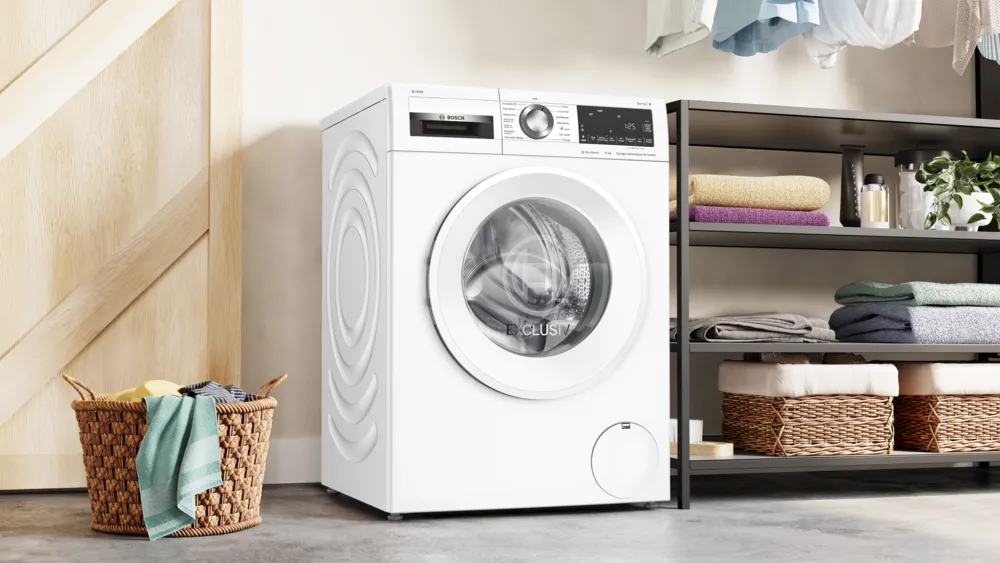 Un lave-linge BOSCH WGG254FAFR se trouve à côté d'un panier à linge, avec des étagères au-dessus soigneusement rangées avec des serviettes pliées et des paniers.