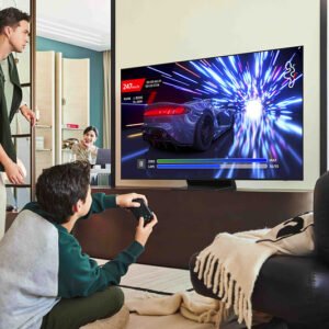 Un garçon joue à un jeu vidéo de course avec une manette sur un téléviseur SAMSUNG TQ55S90F, tandis que deux adultes regardent dans un salon moderne.