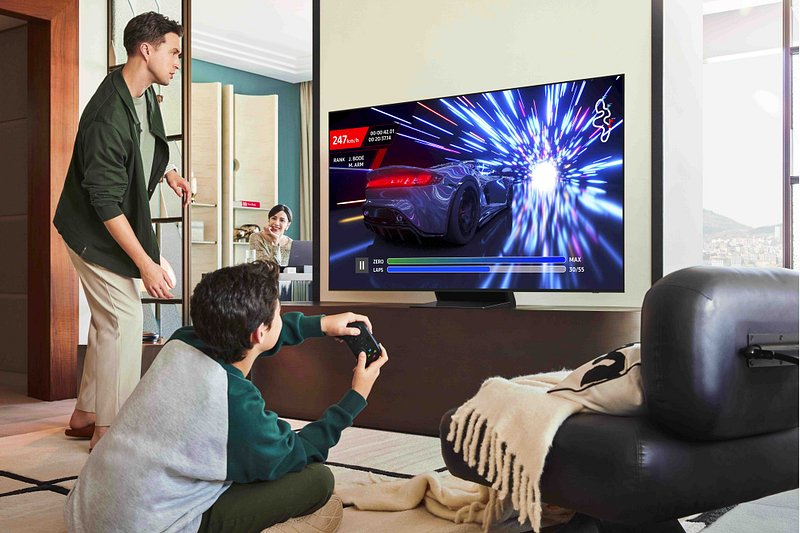 Un garçon joue à un jeu vidéo de course avec une manette sur un téléviseur SAMSUNG TQ55S90F, tandis que deux adultes regardent dans un salon moderne.