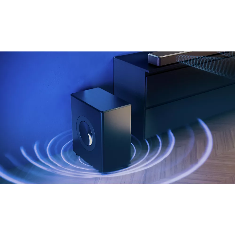 Un caisson de basse PHILIPS TAFW1/10 noir est posé à côté d'un meuble TV sur un sol en bois, contre un mur bleu, et émet des ondes sonores.